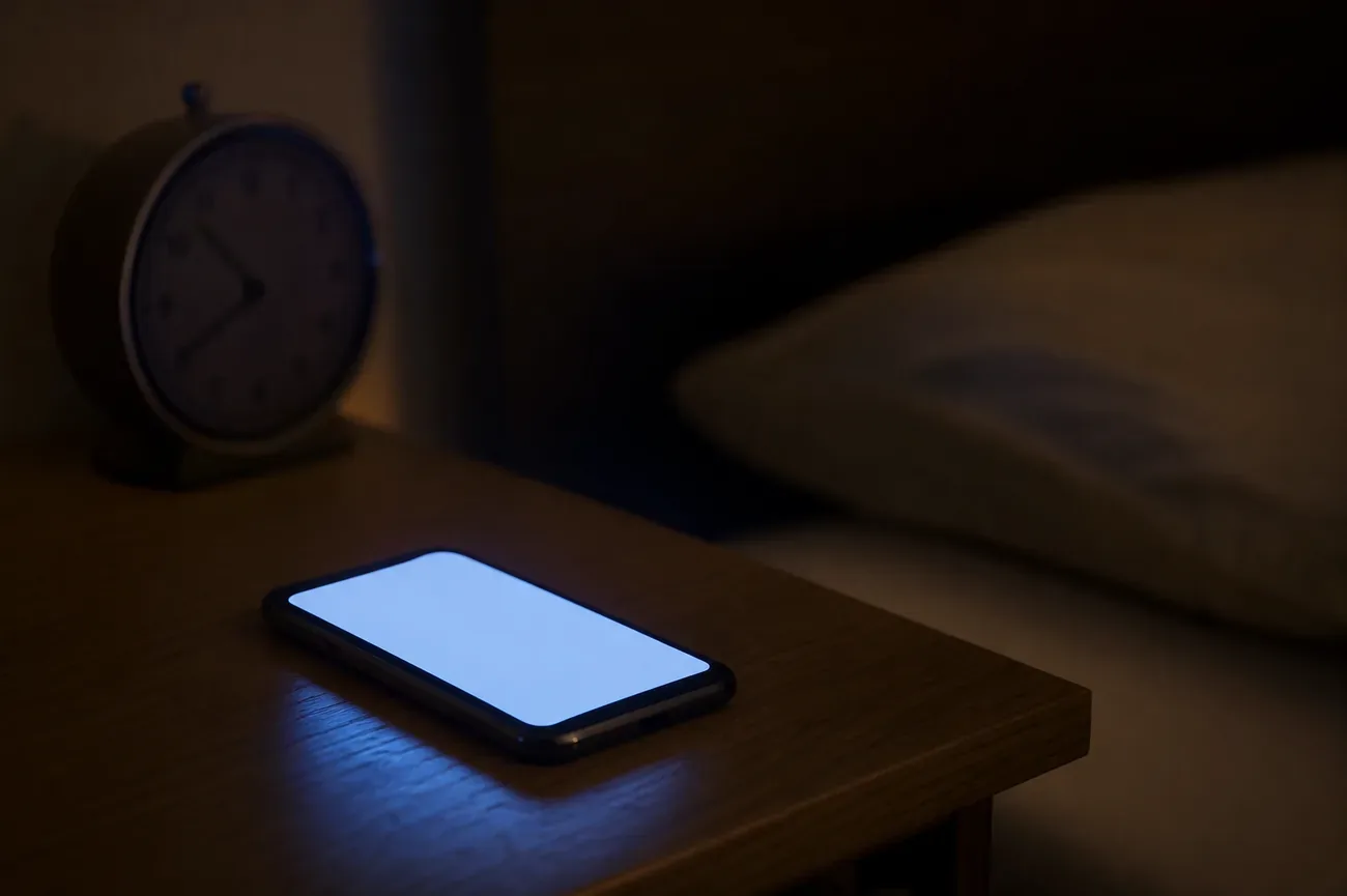 Smartphone glowing soft blue on a wooden nightstand at night, analog alarm clock in soft focus beside it, dimly lit bedroom