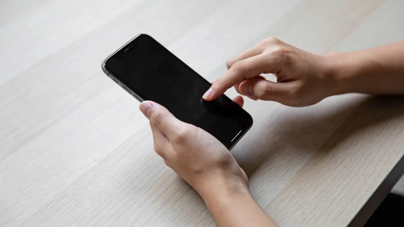 Hands holding a modern smartphone on a light wooden desk, fingertip tapping the screen