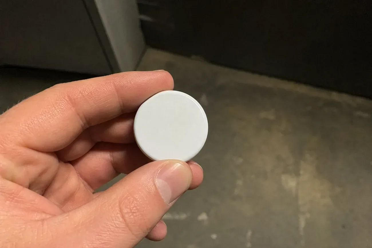A small white disc-shaped tracker held between two fingers against a blurred indoor background, single harsh overhead light, editorial documentary aesthetic