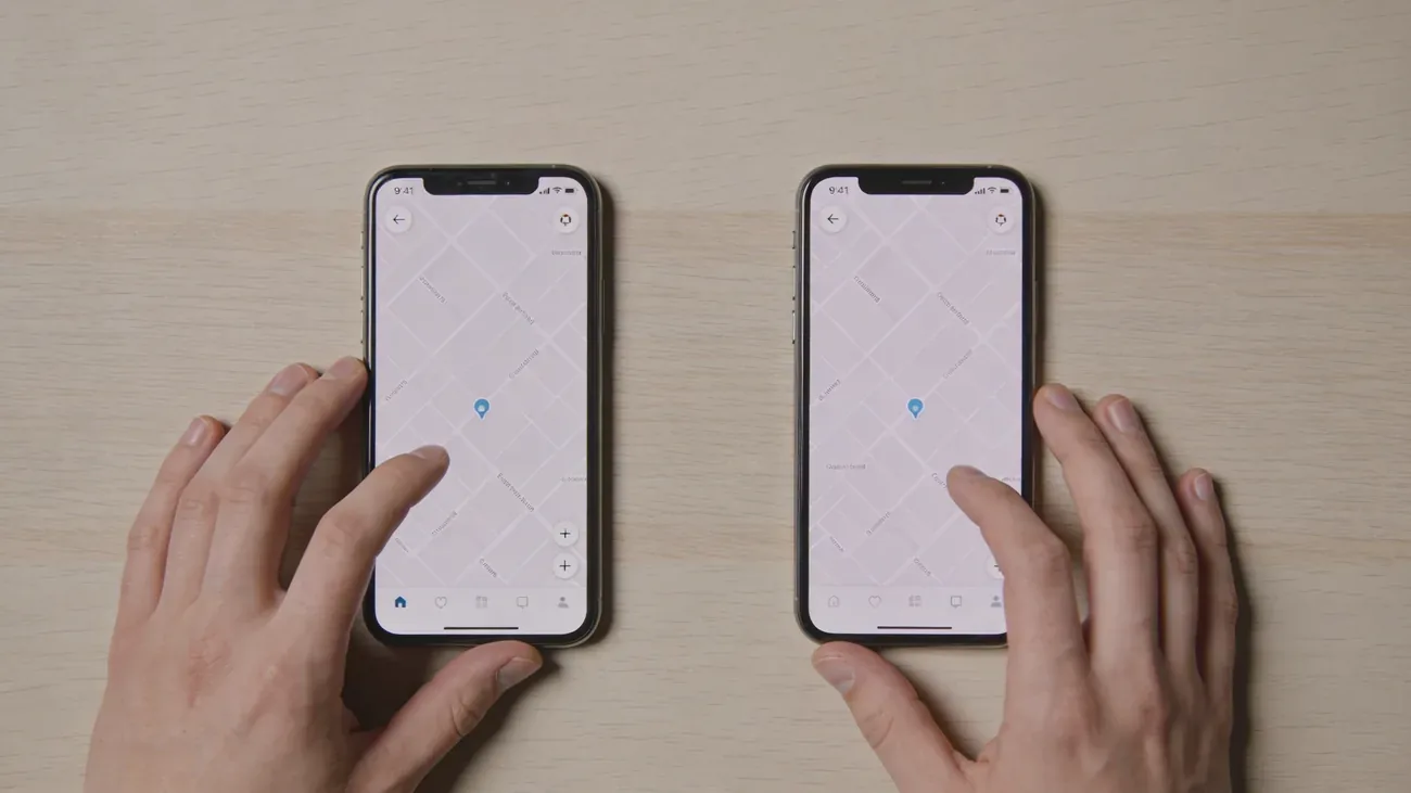 Two smartphones laying side by side on a wooden table, two hands gently touching their own phone, balanced composition
