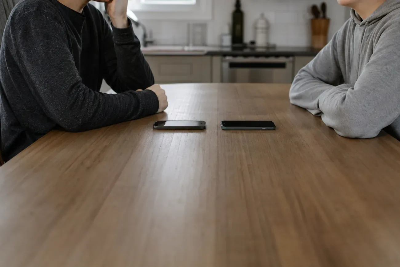 Parent and teenager sitting together at a kitchen table having a calm conversation, phone visible but face-down between them