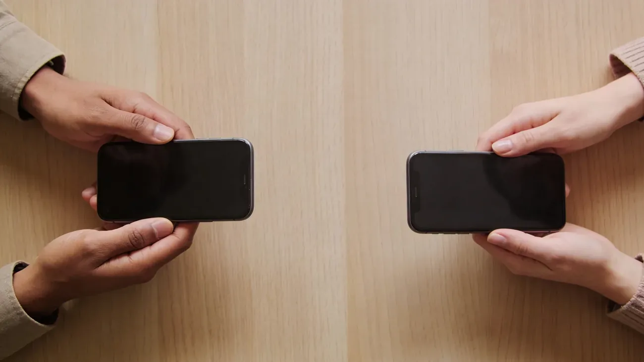 Two pairs of hands meeting on a wooden table, each holding a smartphone with screens off, equal balanced composition